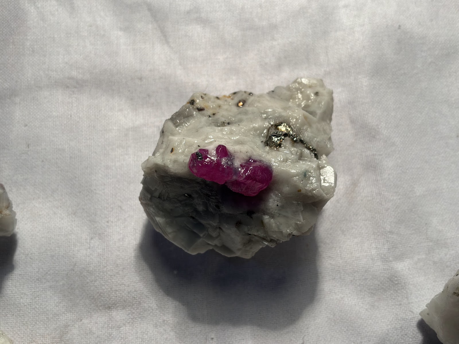 A magenta ruby crystal group sitting proud on a rough block of white calcite marble, with brassy pyrite grains and dark mica flecks in the marble around it, on a white cloth