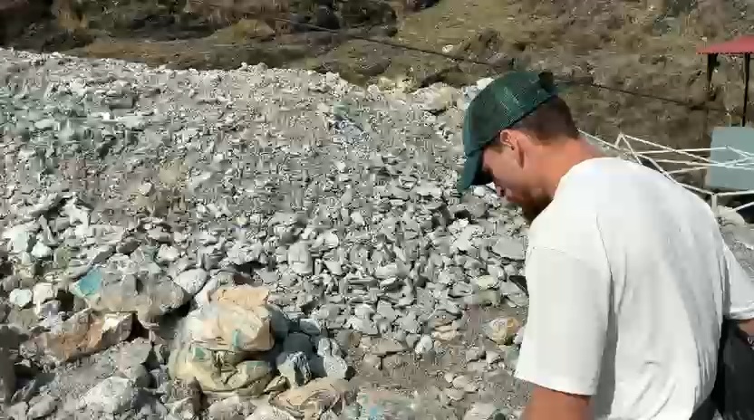 A man in a green cap standing at the edge of a wide spread of broken pale rock, leaning forward to look down at it, with filled sacks at the foot of the slope